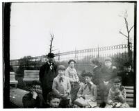 [Group of children with adult male by fence and benches] [graphic].
