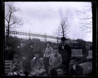 [Group of children with adult male by fence and benches] [graphic].