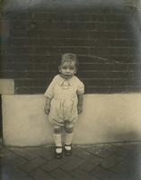 Little boy in playsuit standing in front of brick wall, Philadelphia.