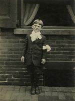 Boy wearing a suit and ribbon bow on arm standing in front of brick house, Philadelphia.