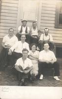 Seven men and one woman sitting on the steps of a wooden house, Philadelphia.