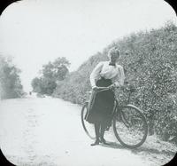 [Woman standing with bicycle in front of hedge near path, Long Island, N.Y.]
