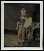 [Portrait of a little boy standing beside a table holding a kite in his left hand.]