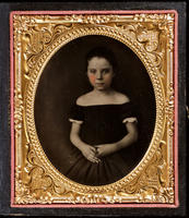 [Unidentified, somber looking, little girl wearing an off the shoulder black dress, her hands folded in her lap.]
