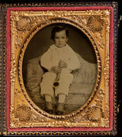 [Portrait of an unidentified boy, dressed in white, sitting on a sofa holding a toy musical instrument.]